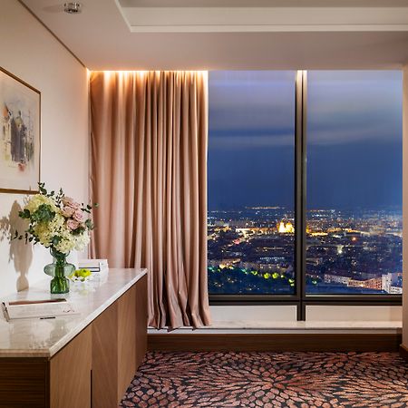 Grand Hotel Millennium Sófia Exterior foto The photo depicts a modern hotel room or a lounge area with a stunning view of a cityscape at night. On the left, there's a wooden console table with a vase of flowers and a bottle of water. A piece of art is framed on the wall above the table. The r