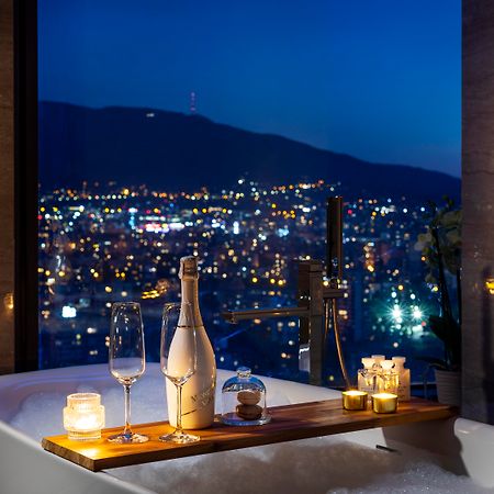 Grand Hotel Millennium Sófia Exterior foto The photo features a luxurious bathtub placed by a large window, showcasing a scenic city view at night. There is a wooden tray resting on the edge of the tub, displaying a bottle of champagne, two glass flutes, and several lit candles, creating an a