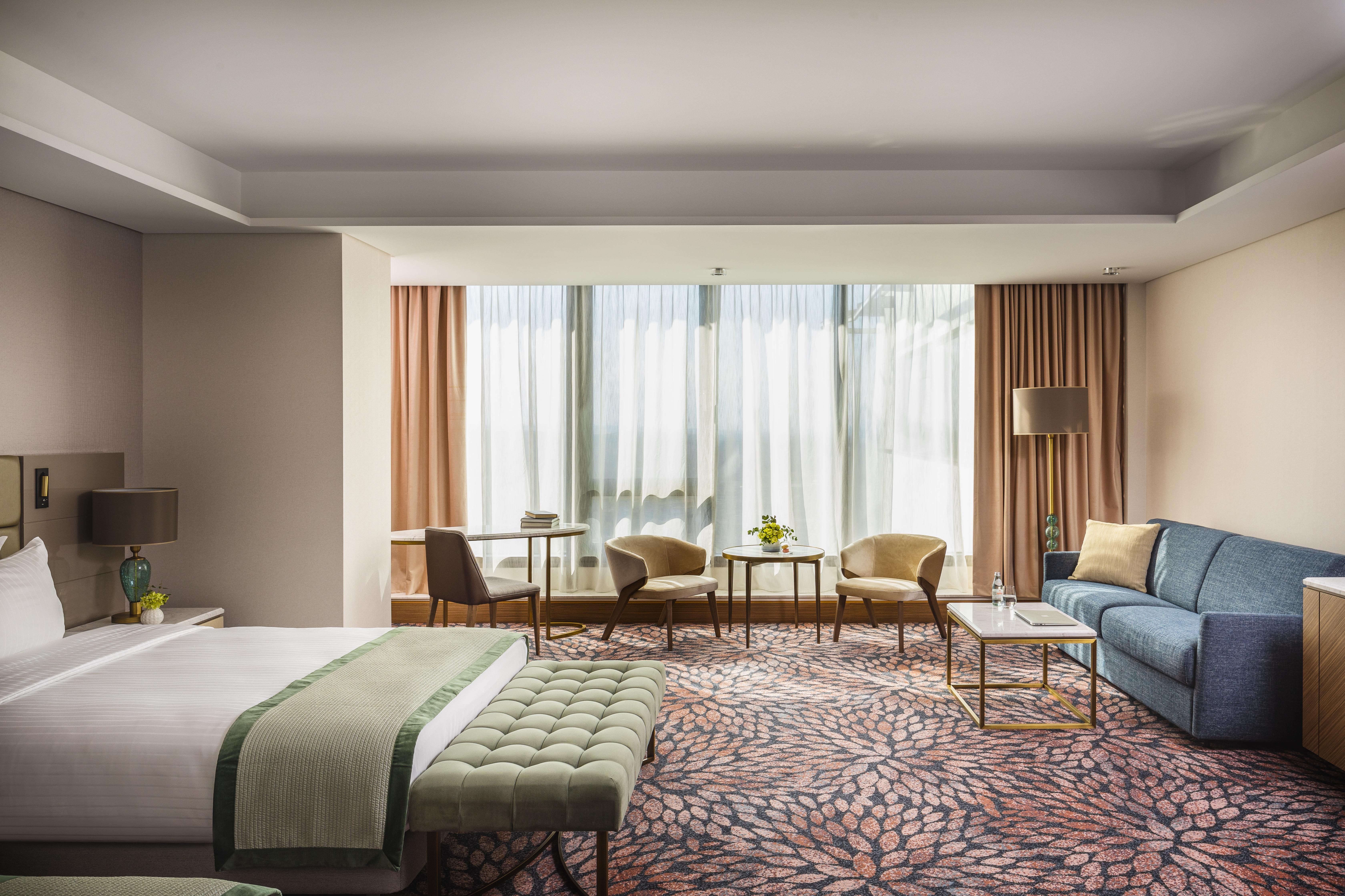 Grand Hotel Millennium Sófia Exterior foto The photo shows a modern hotel room featuring a spacious layout. On one side, there is a large bed with a green throw blanket and a tufted bench at its foot. The room is well-lit by large windows dressed with light curtains, which allow natural light