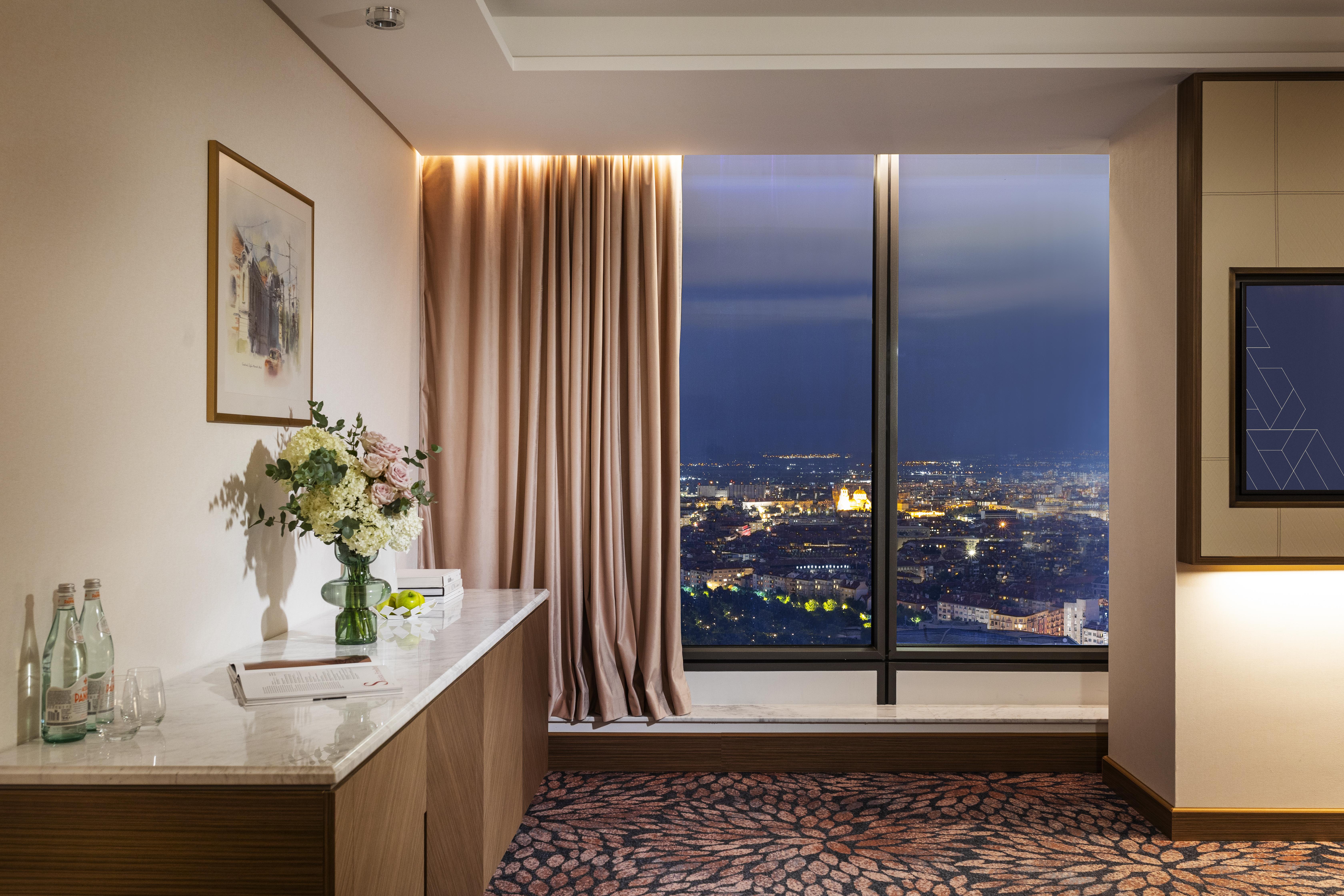Grand Hotel Millennium Sófia Exterior foto The photo depicts a modern hotel room or a lounge area with a stunning view of a cityscape at night. On the left, there's a wooden console table with a vase of flowers and a bottle of water. A piece of art is framed on the wall above the table. The r