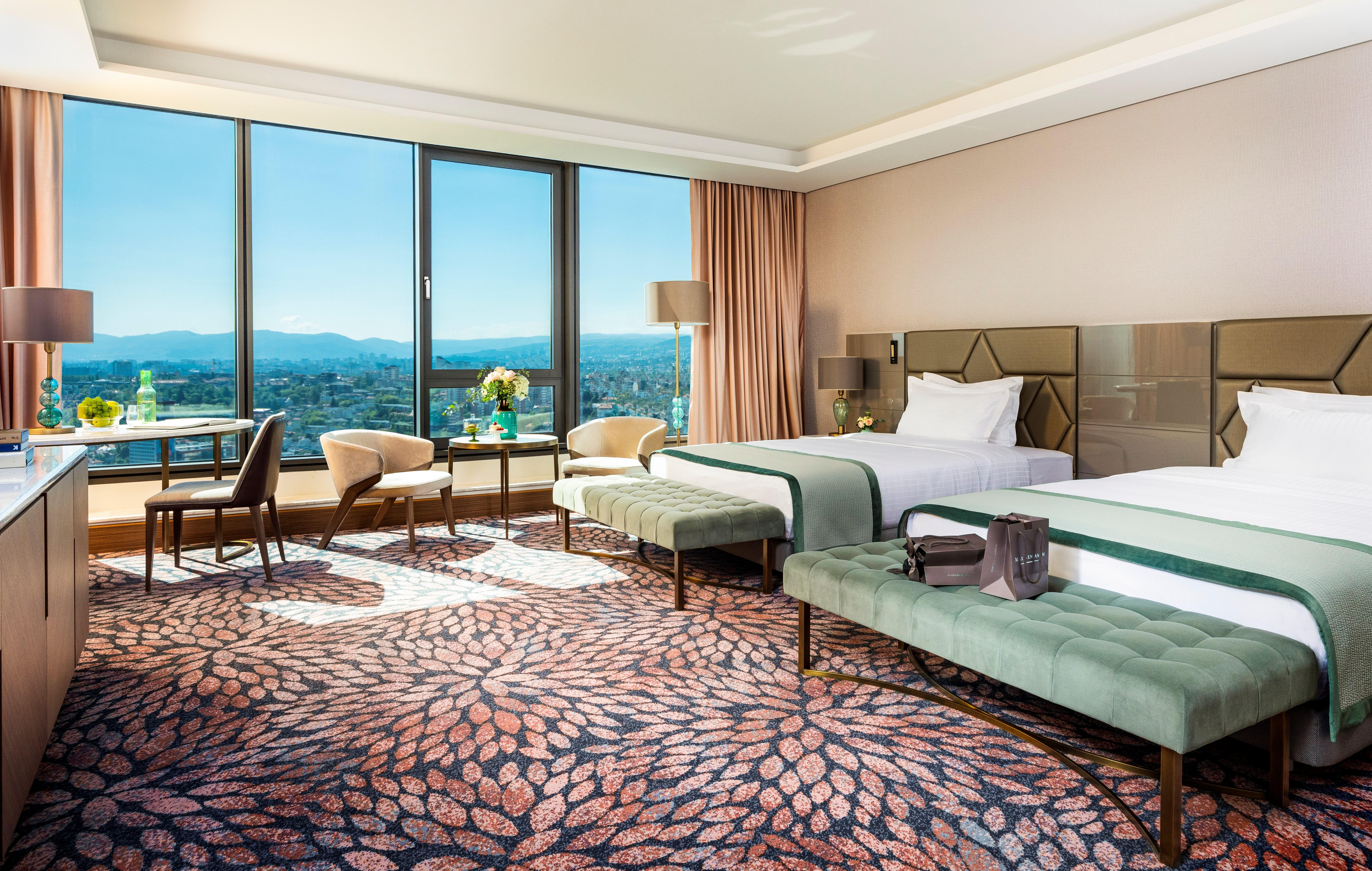 Grand Hotel Millennium Sófia Exterior foto The photo depicts a luxurious hotel room featuring two beds with elegant bedding. The room has large windows that offer a panoramic view of the city and mountains in the background. There is a decorative area rug with a floral pattern covering the fl