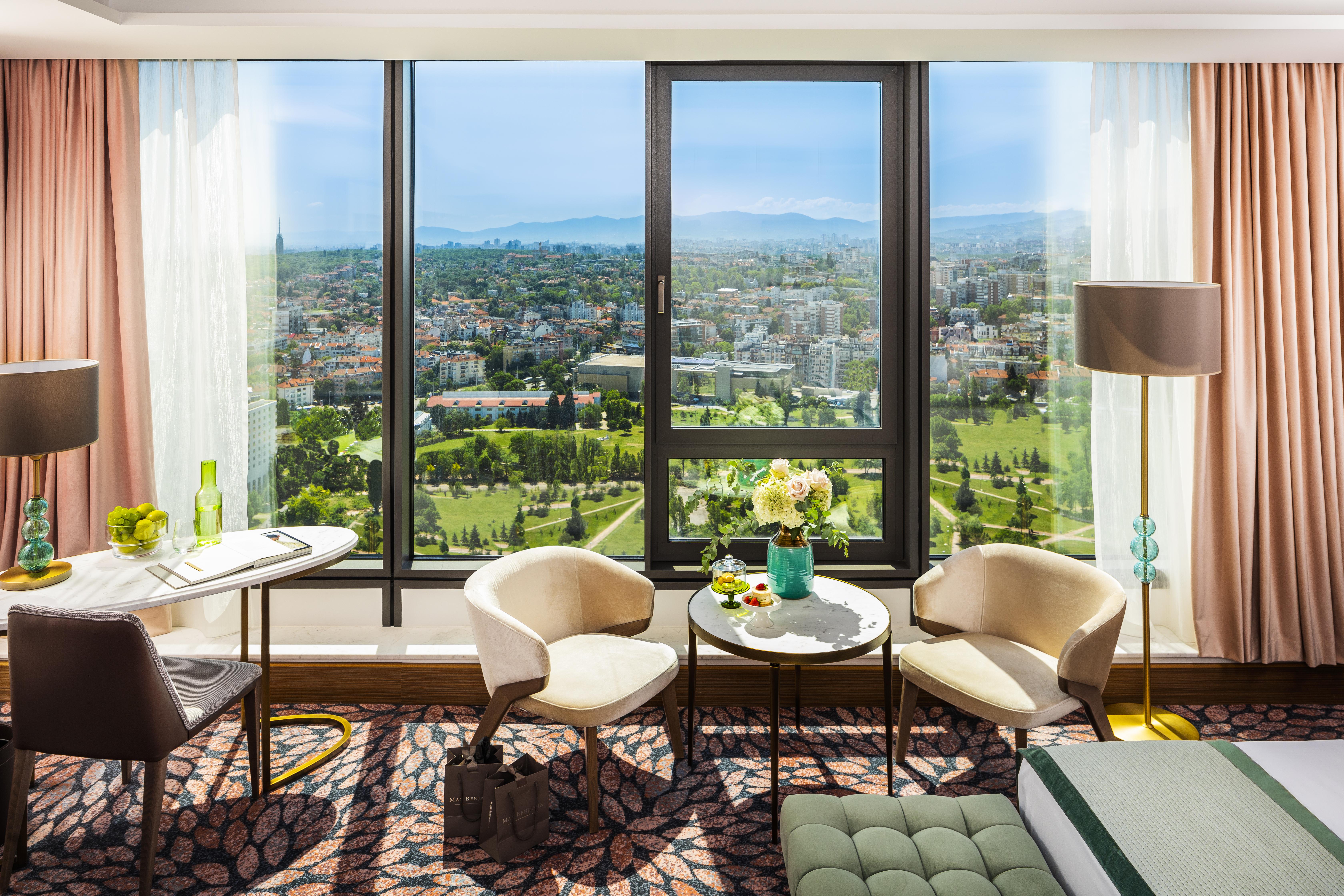 Grand Hotel Millennium Sófia Exterior foto The photo shows a stylish hotel room with large windows providing a beautiful view of a cityscape and landscape beyond. There are two modern chairs and a small round table set near the windows, accompanied by a vase of fresh flowers. The room feature
