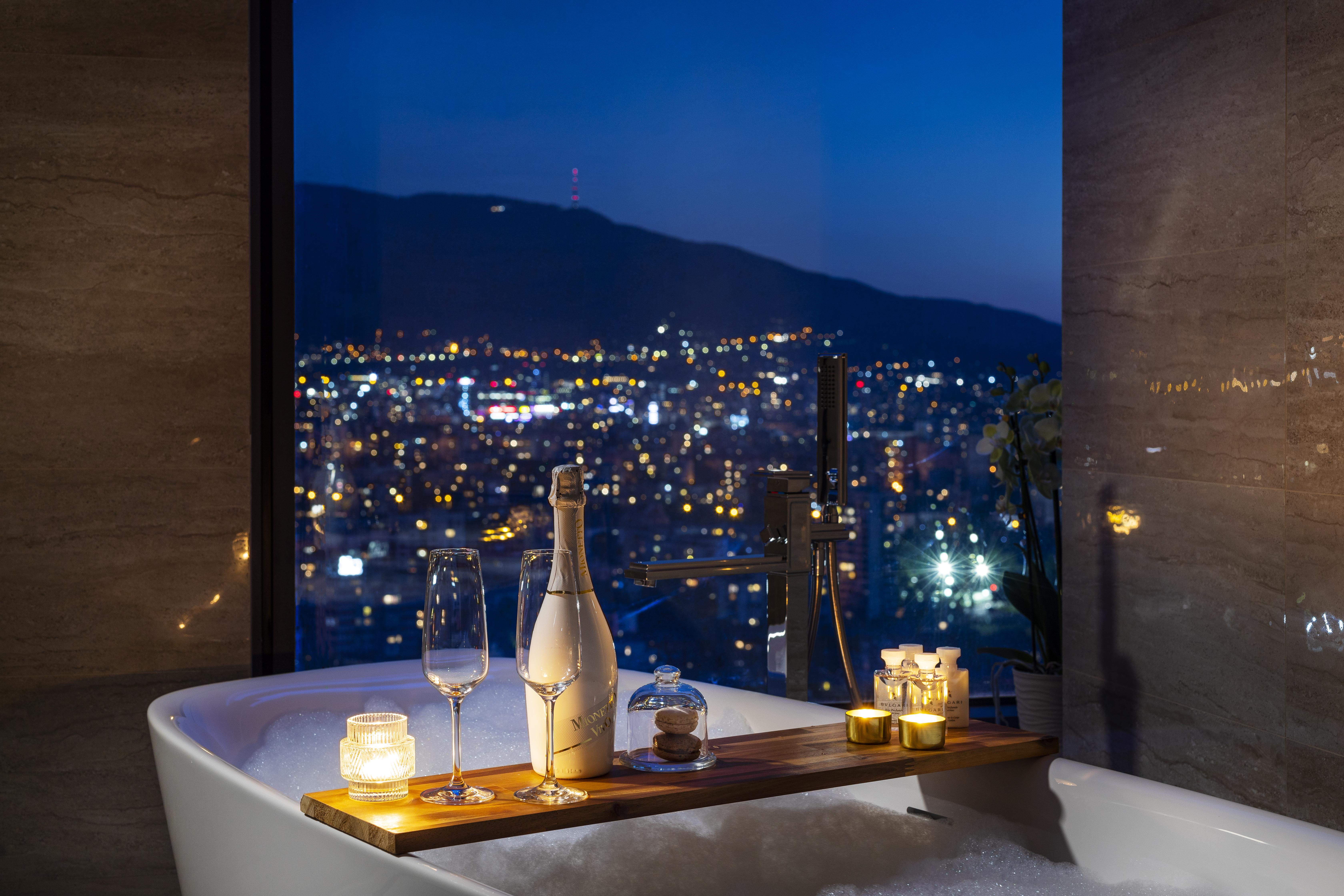 Grand Hotel Millennium Sófia Exterior foto The photo features a luxurious bathtub placed by a large window, showcasing a scenic city view at night. There is a wooden tray resting on the edge of the tub, displaying a bottle of champagne, two glass flutes, and several lit candles, creating an a