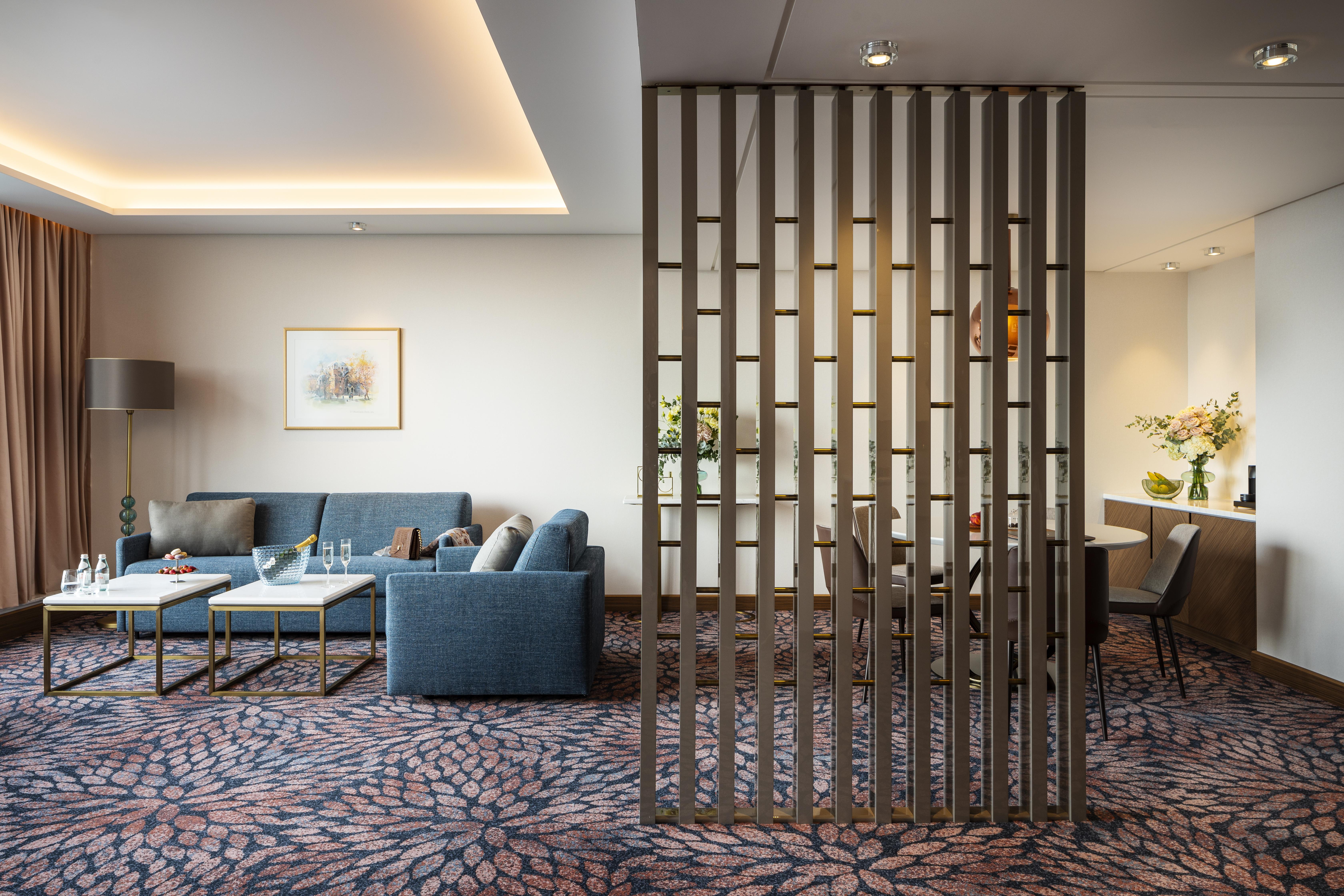 Grand Hotel Millennium Sófia Exterior foto The photo shows a modern living space with a comfortable seating area featuring a blue couch and two coffee tables. There is a decorative partition made of vertical wooden slats, which separates this sitting area from a dining space. The dining area 