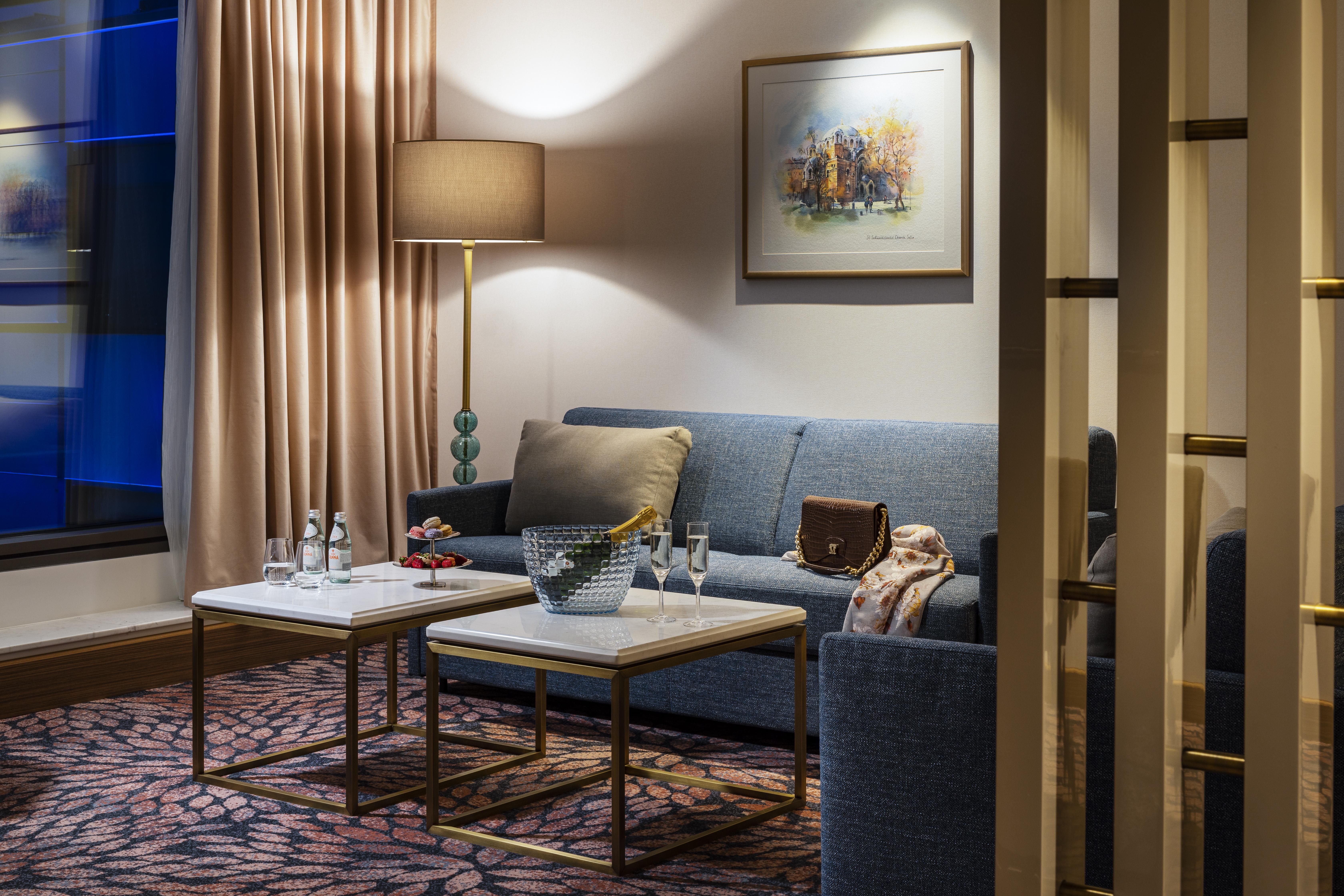 Grand Hotel Millennium Sófia Exterior foto The photo shows a cozy and elegantly decorated living space. It features a blue sofa against a wall, complemented by a painting above it. There are two small marble-top coffee tables with gold frames, one of which holds decorative items and glasses. 