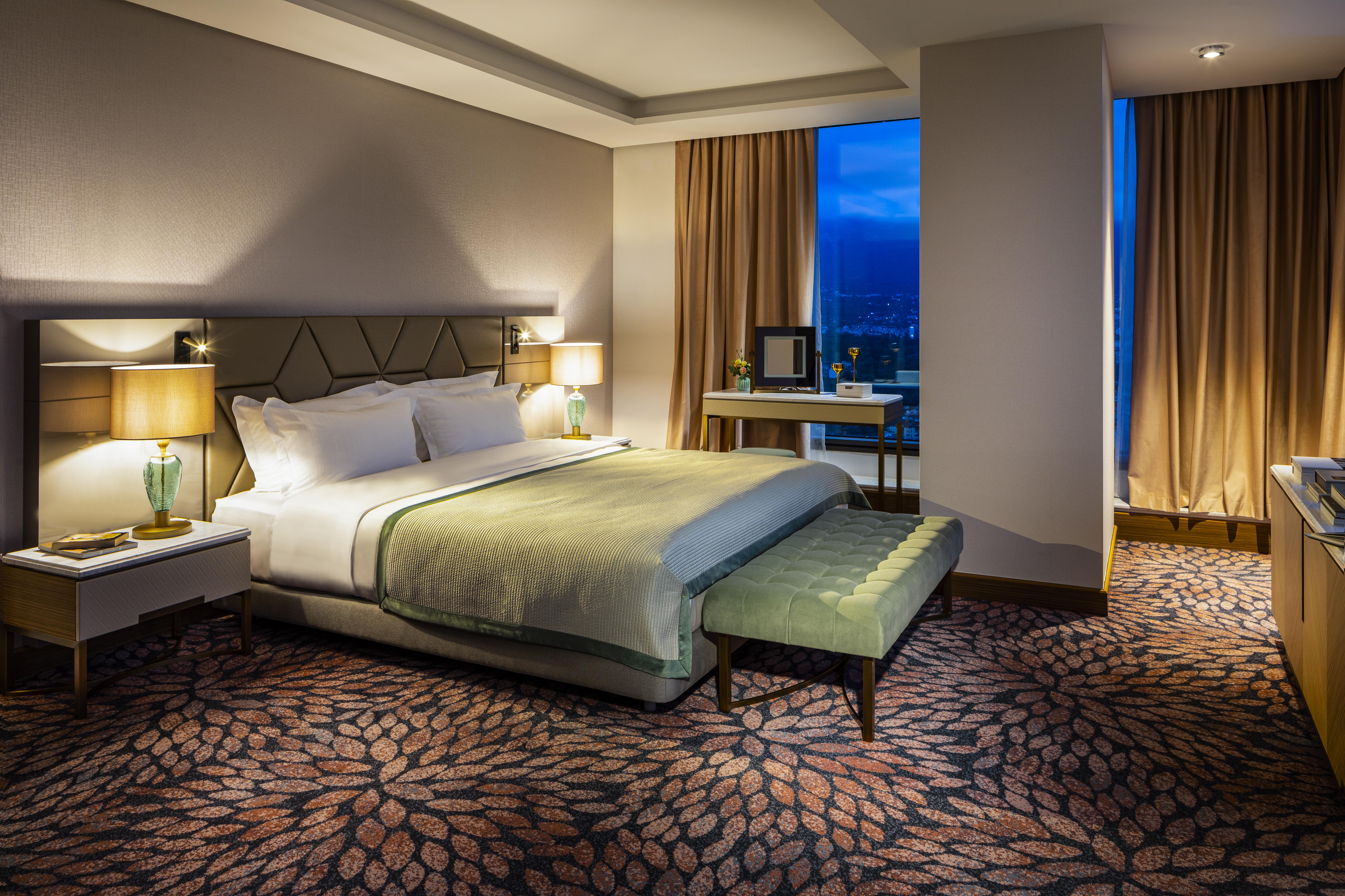 Grand Hotel Millennium Sófia Exterior foto The photo shows a modern hotel room featuring a spacious design. It has a large bed with a light green throw blanket positioned in the center. On either side of the bed, there are stylish bedside tables with lamps. 

The floor has a patterned carpet,
