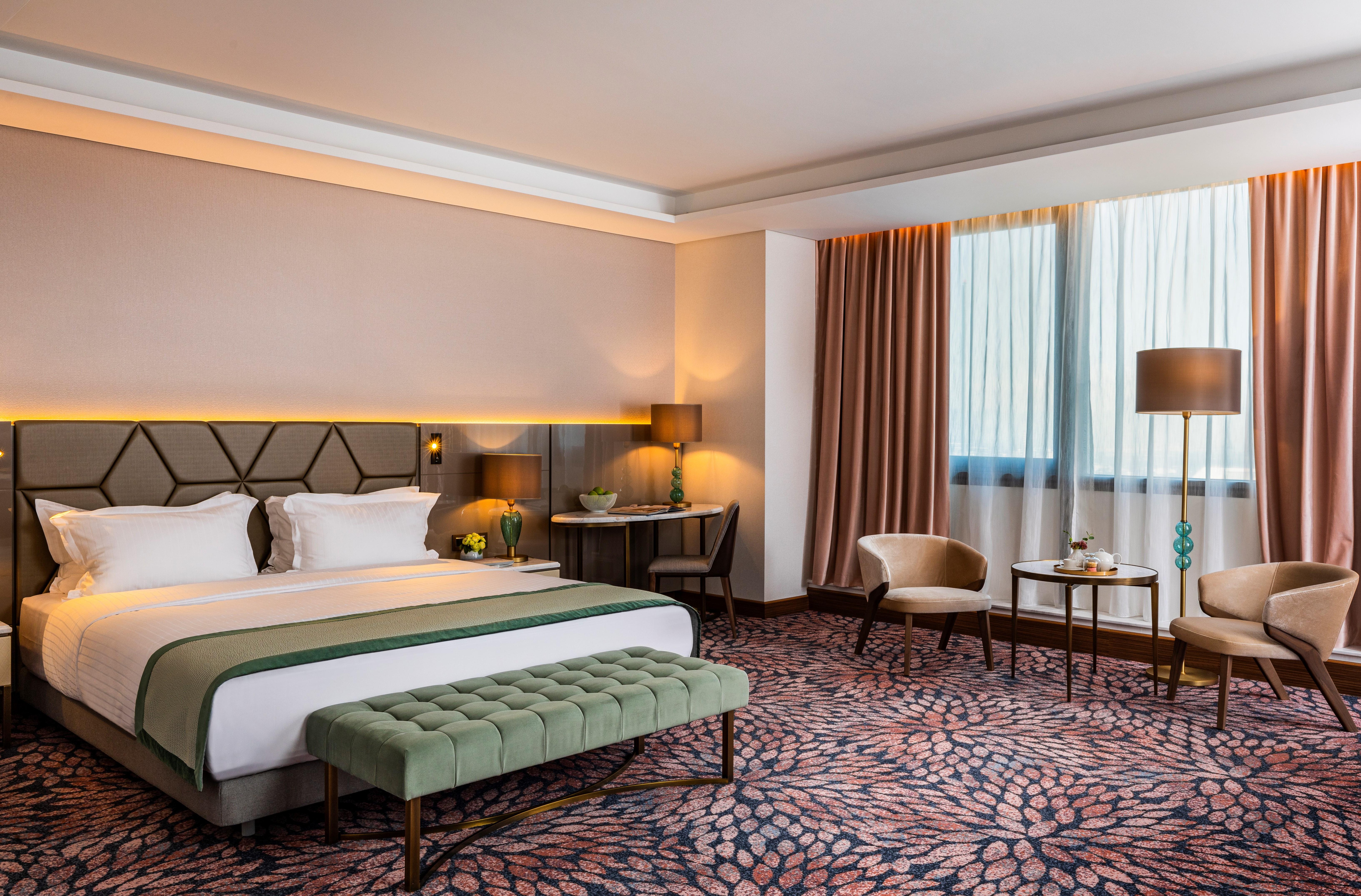 Grand Hotel Millennium Sófia Exterior foto The photo shows a modern hotel room. It features a large bed with a headboard, topped with two white pillows and a green throw blanket. In front of the bed, there is a stylish green bench. On either side of the bed, there are bedside tables with lamp