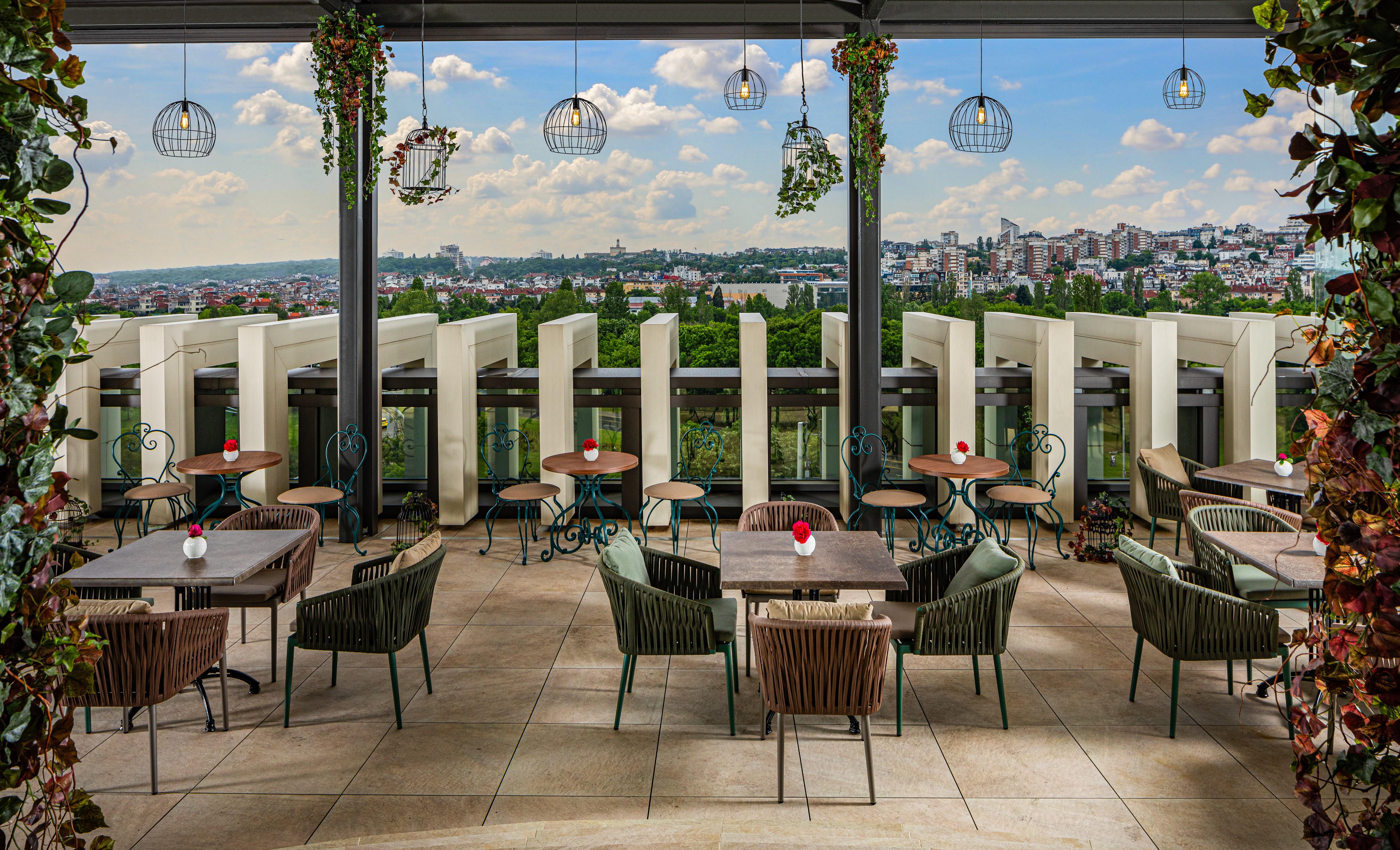 Grand Hotel Millennium Sófia Exterior foto The photo shows a stylish outdoor restaurant or café setting. There are several tables arranged on a tiled patio, each with chairs, some made of woven materials. Centerpieces with small red flowers adorn each table. The space is partially shaded by o