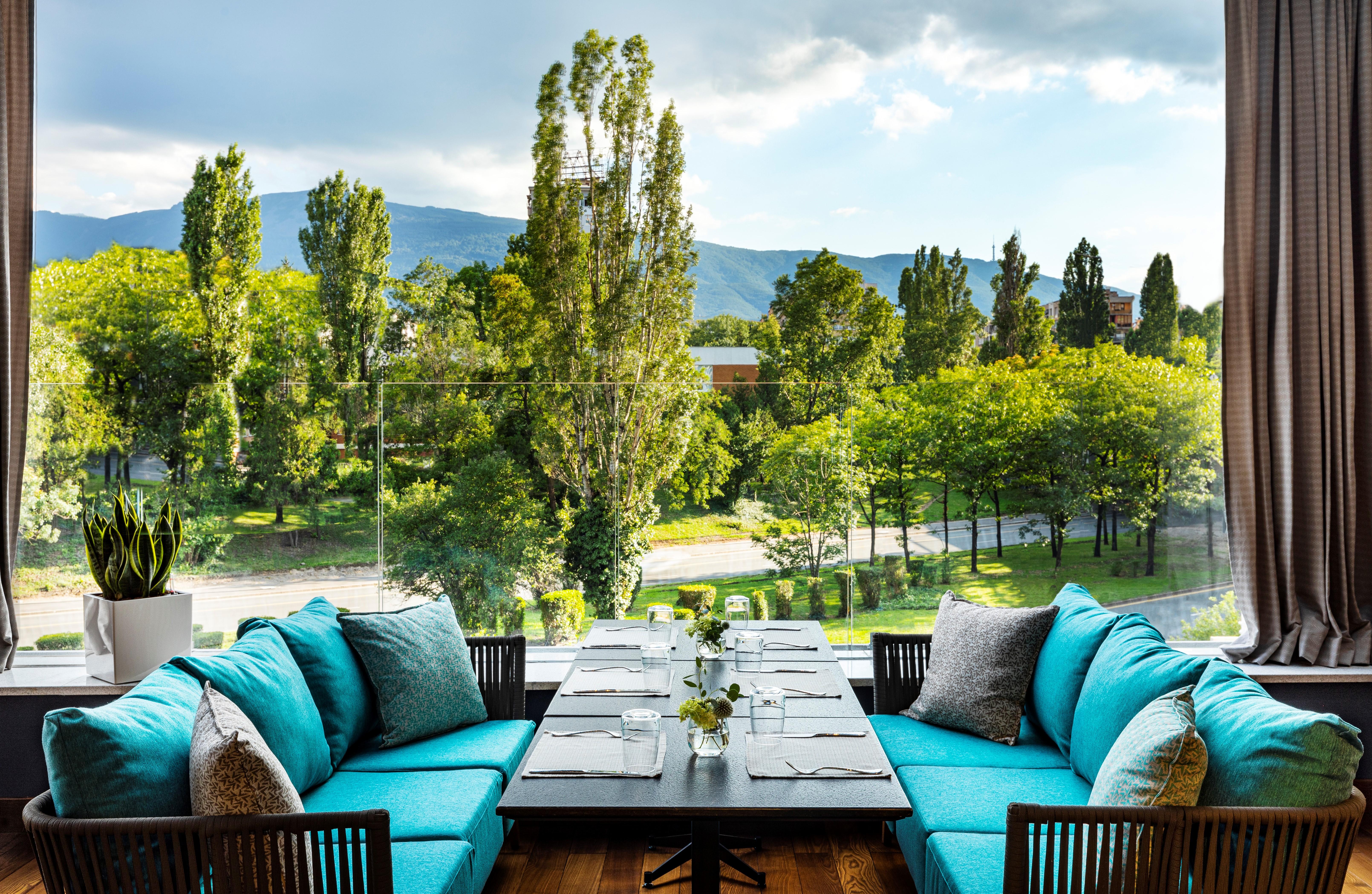 Grand Hotel Millennium Sófia Exterior foto The photo shows a bright and inviting dining area with large windows that overlook a lush green landscape. There are several trees in the background, along with a view of distant mountains under a partly cloudy sky. The seating consists of stylish tu