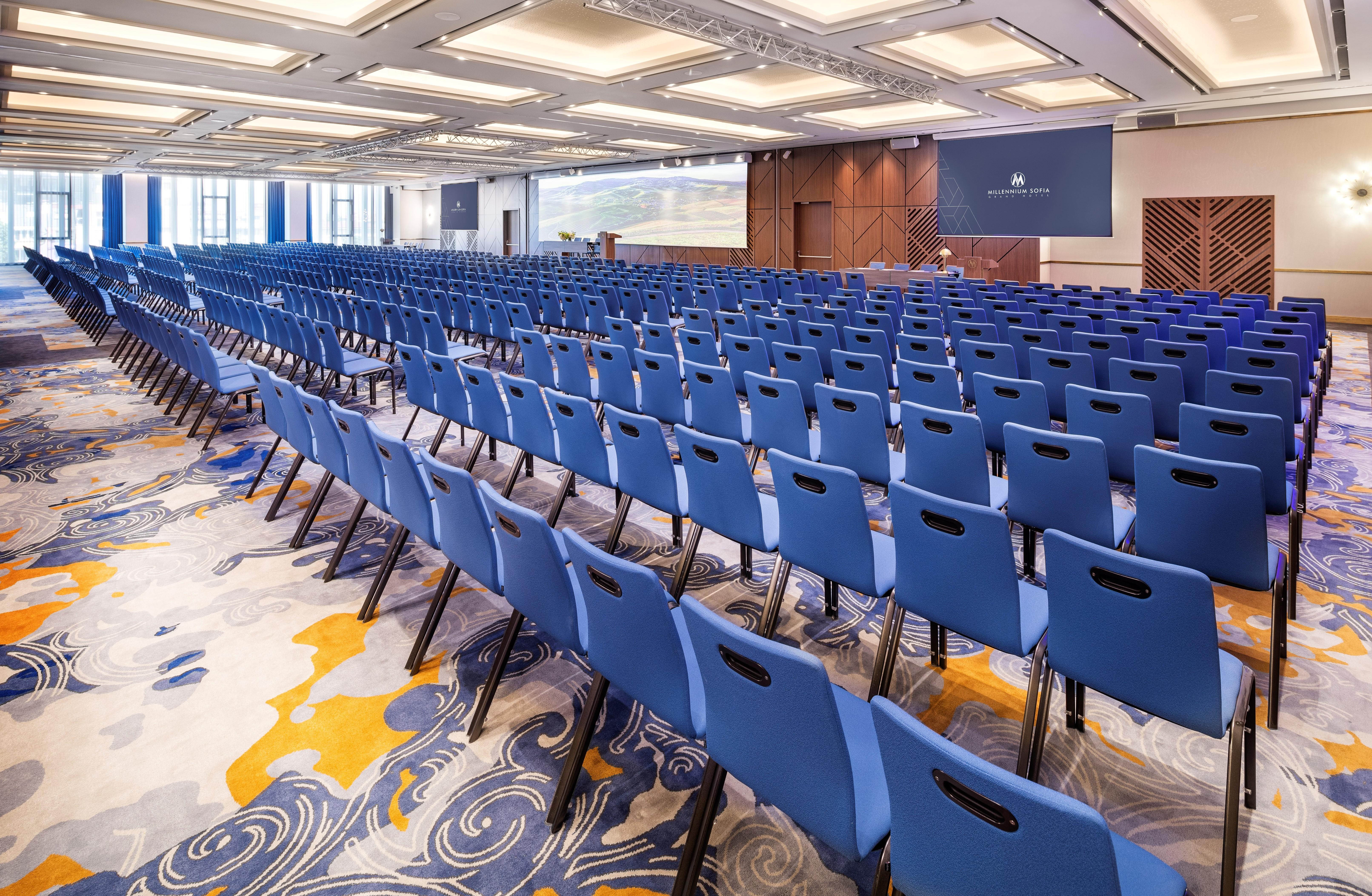 Grand Hotel Millennium Sófia Exterior foto The photo shows a large conference or meeting room set up for an event. There are numerous blue chairs arranged in rows, creating a seating area for attendees. The floor features a decorative pattern, and the room has a modern design with ample light