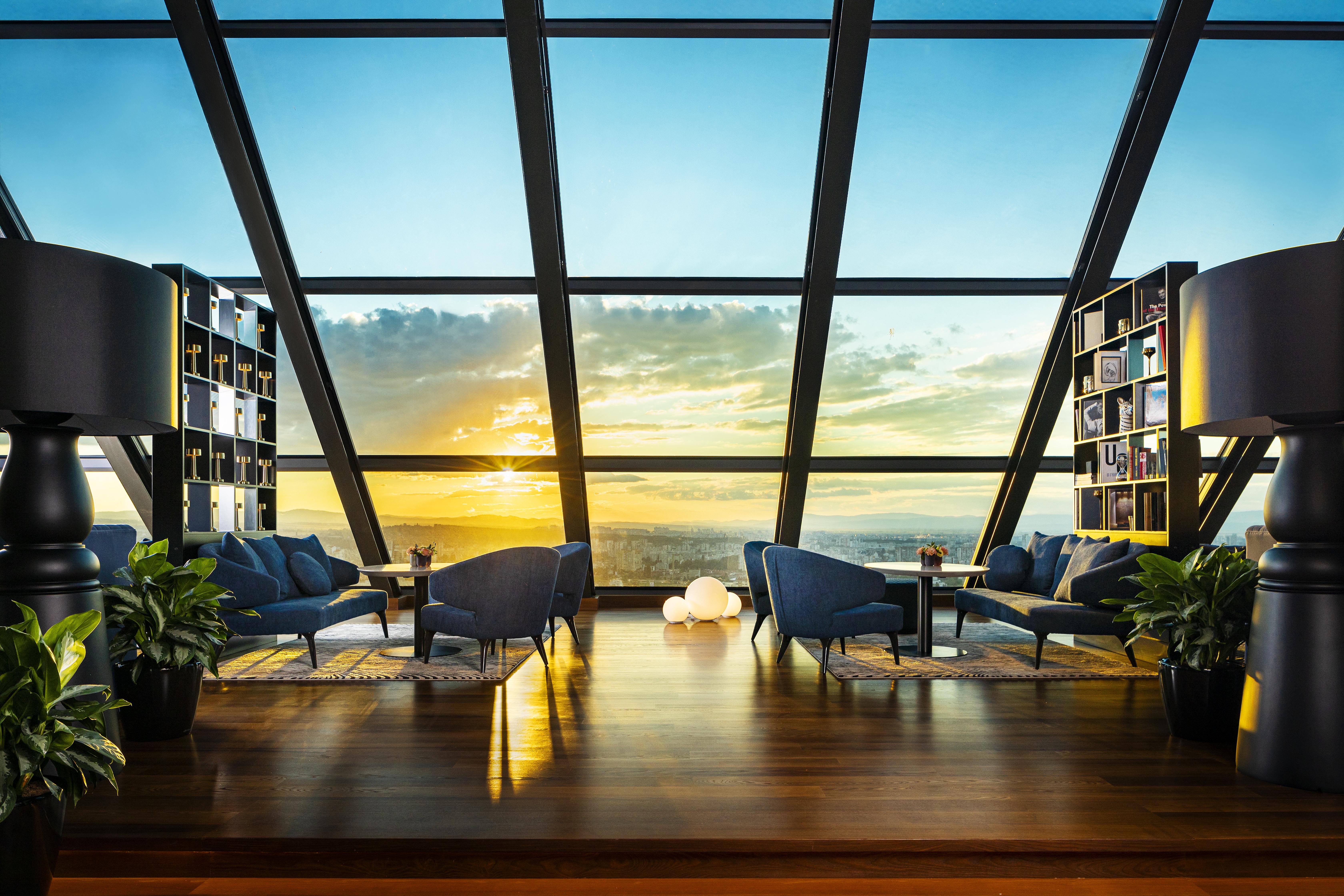 Grand Hotel Millennium Sófia Exterior foto The photo depicts a modern, stylish interior of a high-rise space with a panoramic view through large glass windows. The scene features a cozy seating area with several blue armchairs arranged around a low table, creating a comfortable atmosphere. Th