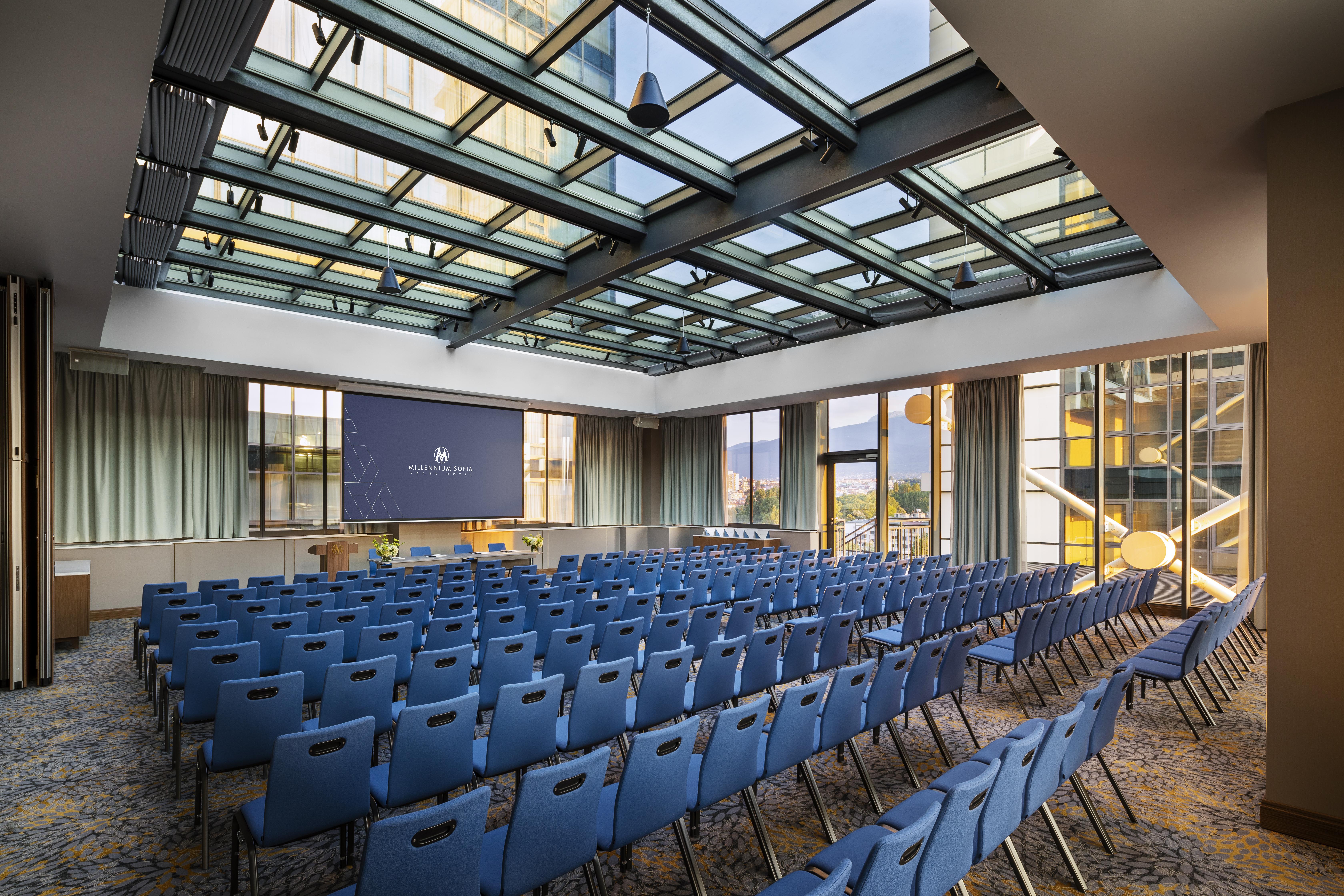 Grand Hotel Millennium Sófia Exterior foto The photo shows a large conference room or meeting space. It features rows of blue chairs arranged in a theater style, facing a large screen at the front of the room. The room is well-lit, with a glass ceiling allowing natural light to filter in. The