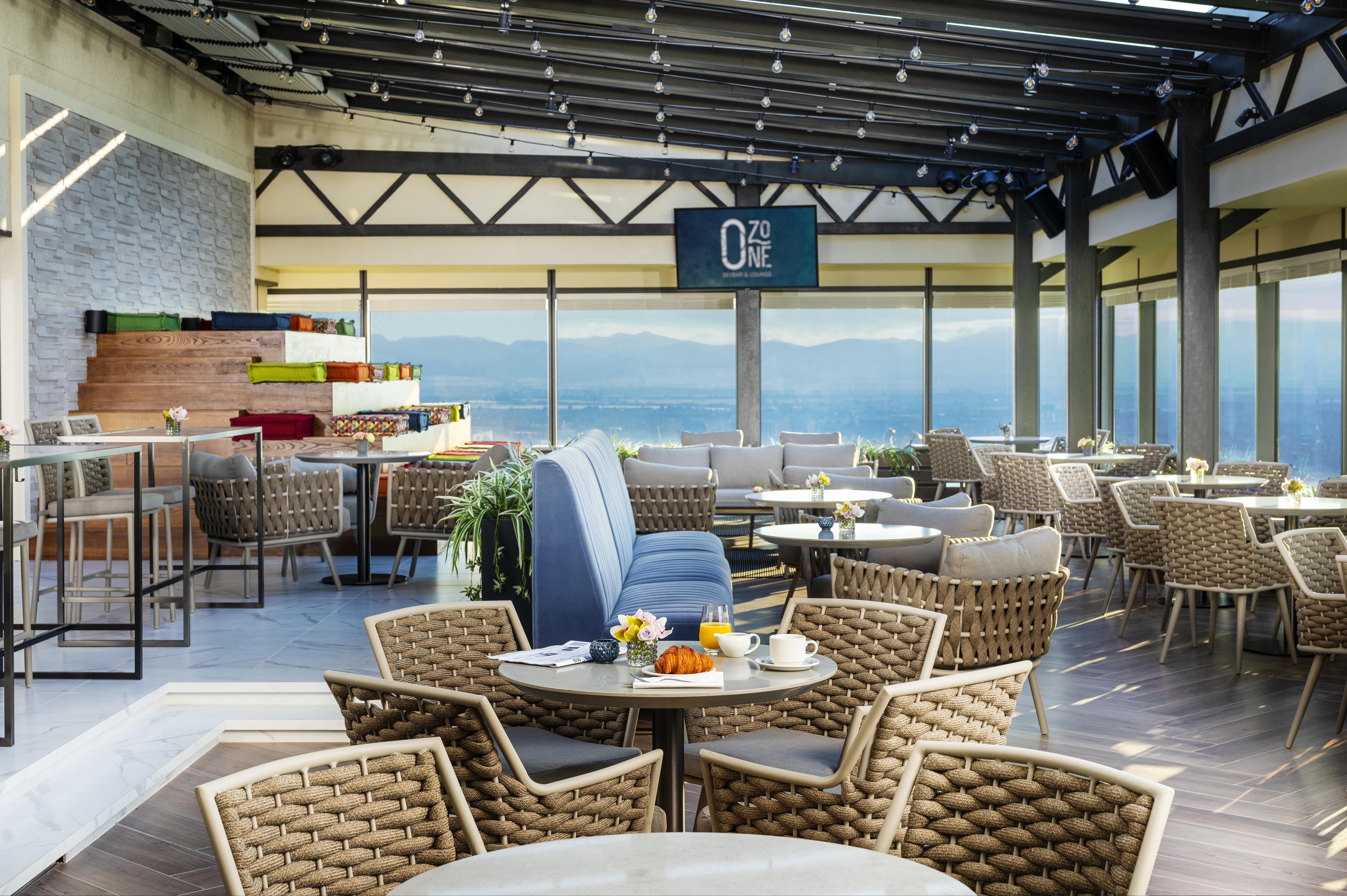 Grand Hotel Millennium Sófia Exterior foto The photo shows a modern restaurant or café with a spacious interior. It features a variety of seating arrangements, including round tables surrounded by wicker chairs and lounges. Large windows allow for a panoramic view, which likely includes sceni