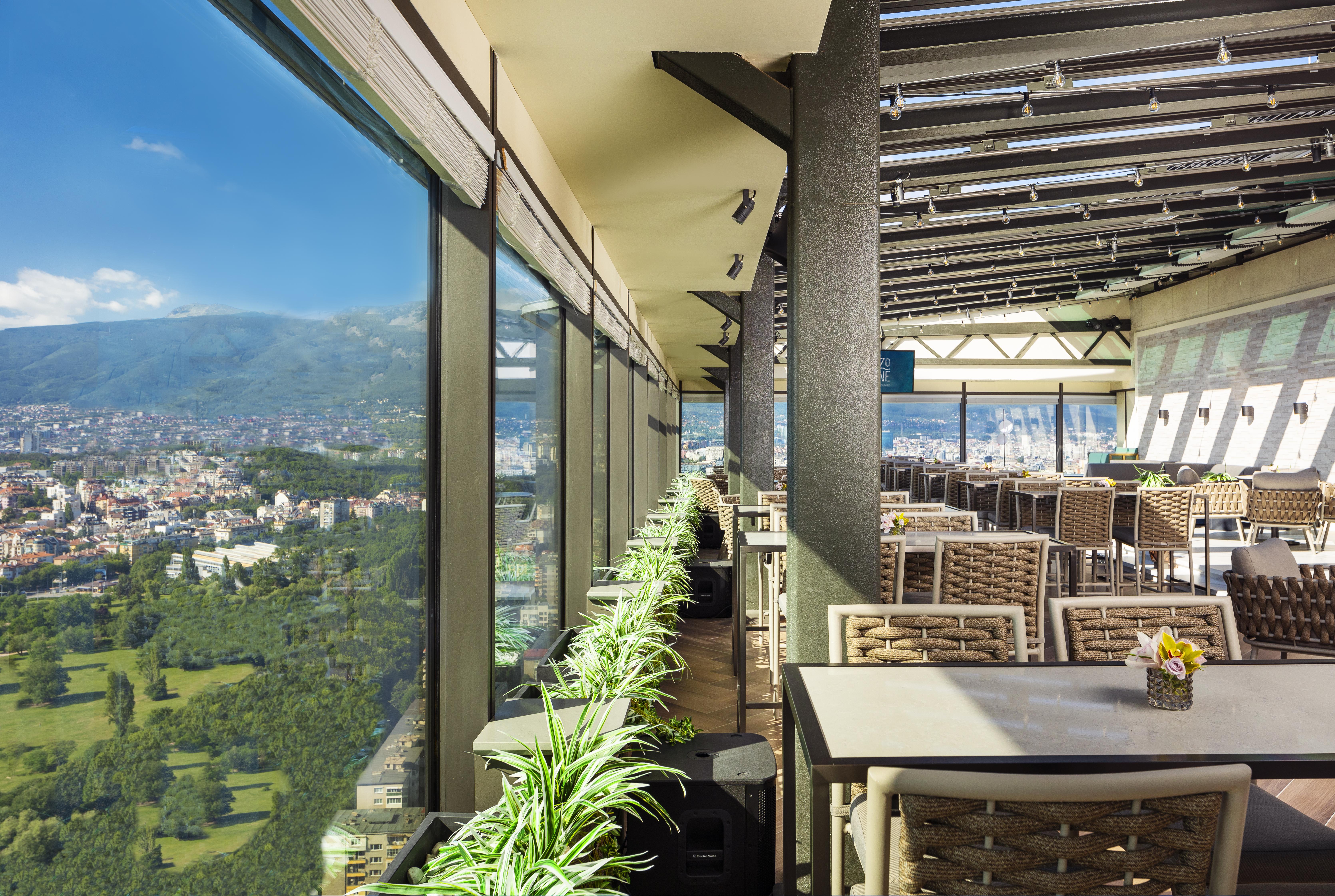 Grand Hotel Millennium Sófia Exterior foto The photo shows a modern, spacious restaurant with large glass windows offering a panoramic view of a city and surrounding landscape. The interior features contemporary furniture, including stylish tables and chairs. There are potted plants along the